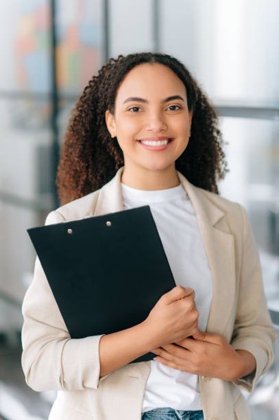 Vertical portrait of a positive beautiful successful brazilian or hispanic business lady, corporate ceo, dressed in elegant clothes, standing in a modern creative office, looks at camera, smiling